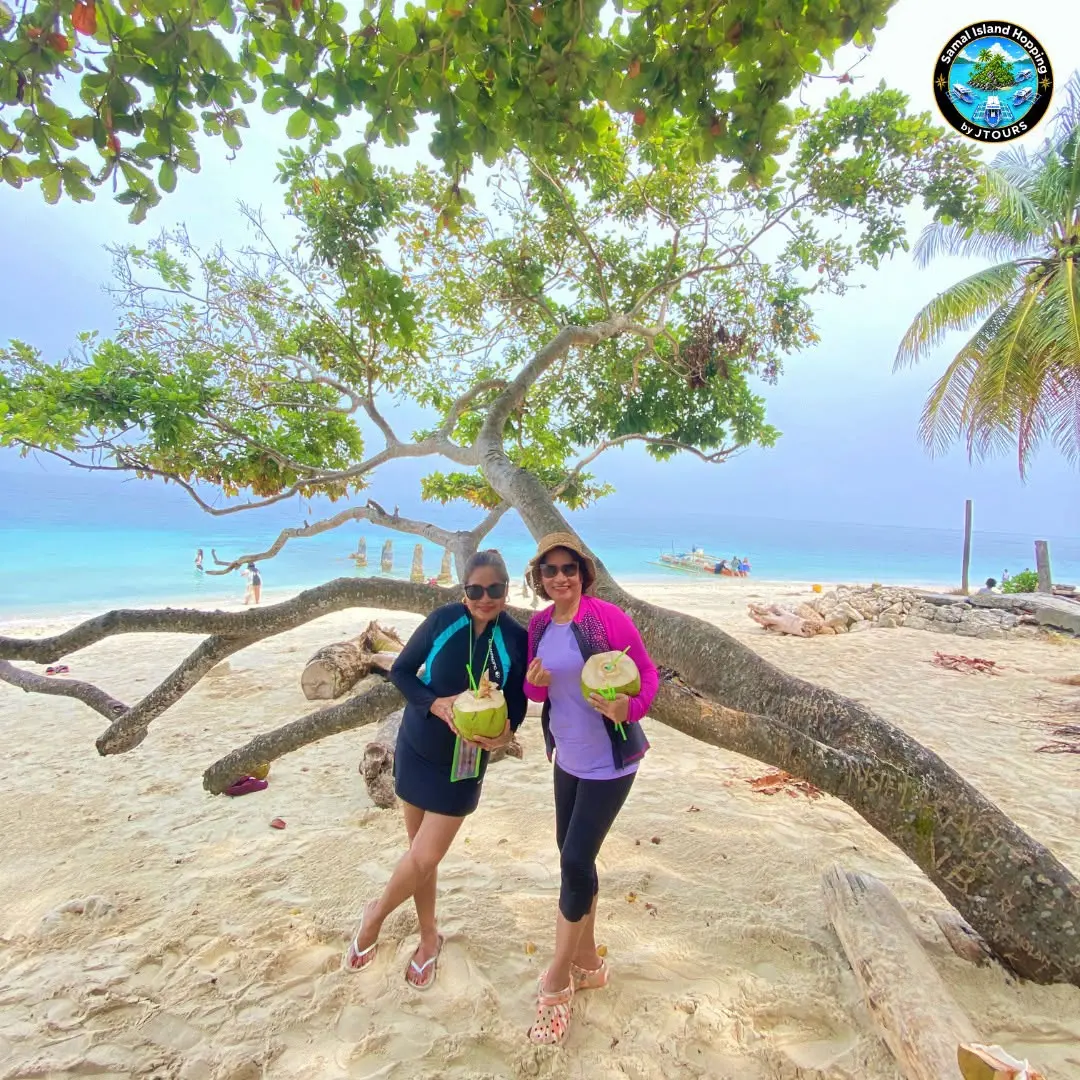 Two guests holding coconuts under a leaning tree on white sand